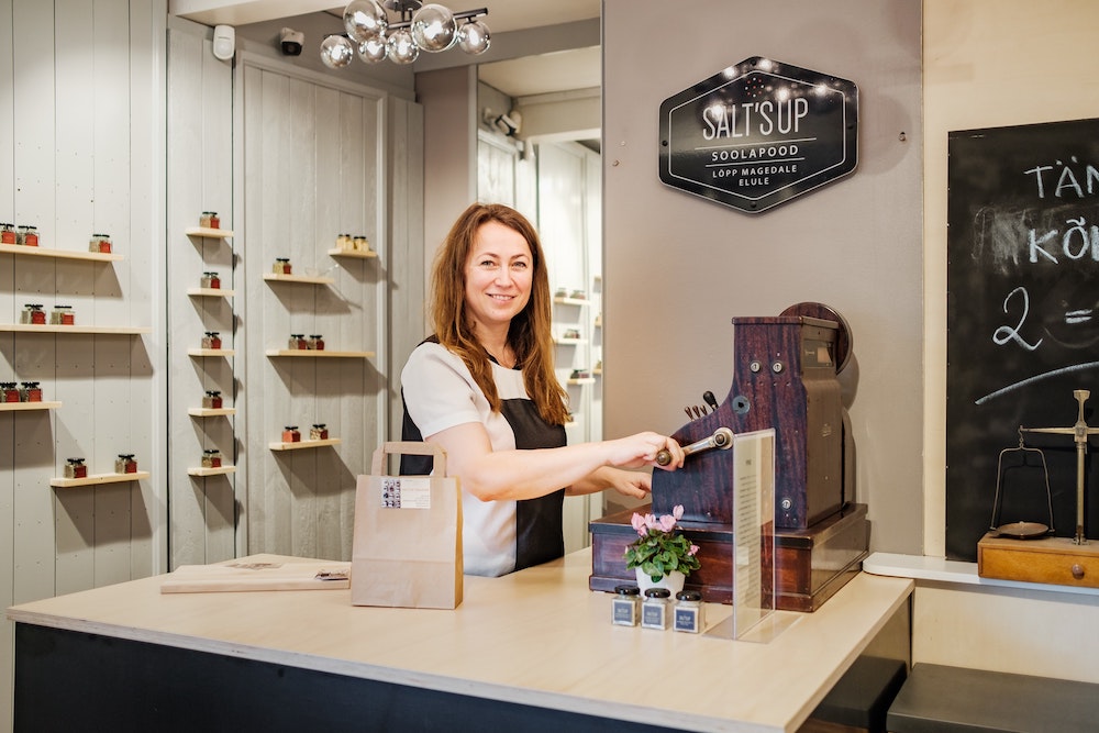 A business owner behind a cash register with a branded shop sign in the background.