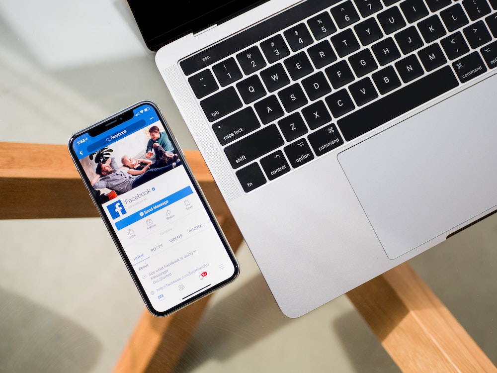 A laptop keyboard on a glass desk, with a phone beside it showing Facebook.