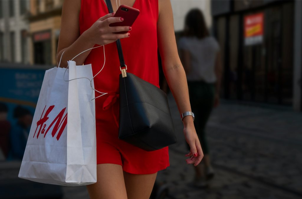 A woman walking in a city street holding an H&M shopping bag and looking at her phone.