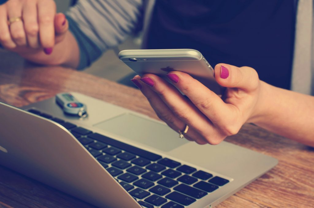 Close-up of hands holding a smartphone above an open laptop on a wooden desk.