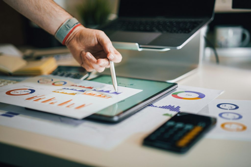 A person reviews colorful survey charts and graphs on paper while using a stylus on a tablet, with a laptop and phone on the desk
