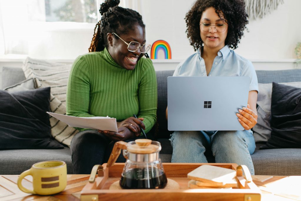 Two people sitting on a couch smiling while reviewing something together on a laptop and printed papers, with coffee on a tray in the foreground.