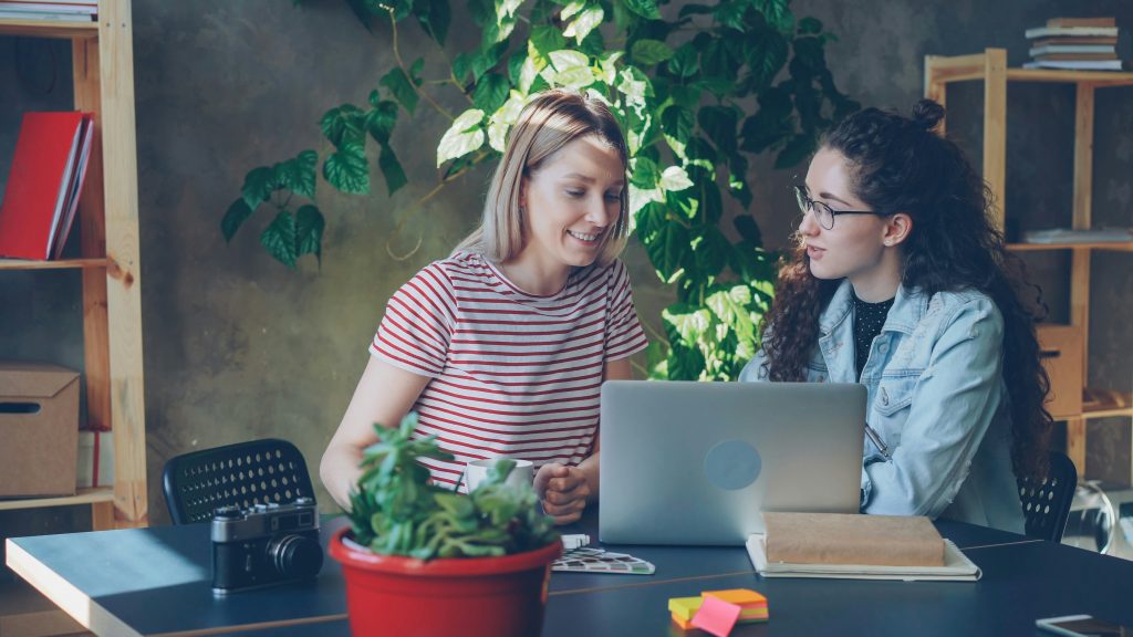 Two colleagues review something on a laptop at a desk in a bright office with leafy plants